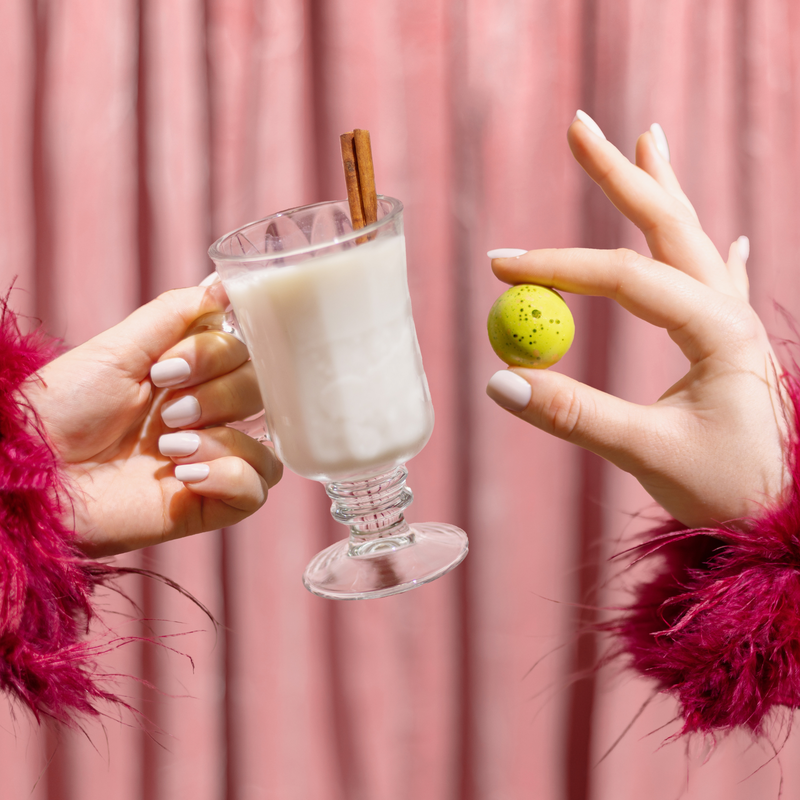 One hand holding a light green Eggnog chocolate beside another hand holding a clear glass of eggnog with a cinnamon stick. The chocolate and the glass of eggnog are held up as if to cheers one another to a great holiday season.